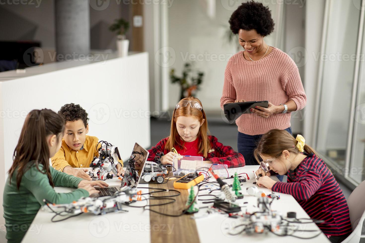 crianças felizes com sua professora de ciências afro-americana com laptop programando brinquedos elétricos e robôs na sala de aula de robótica foto