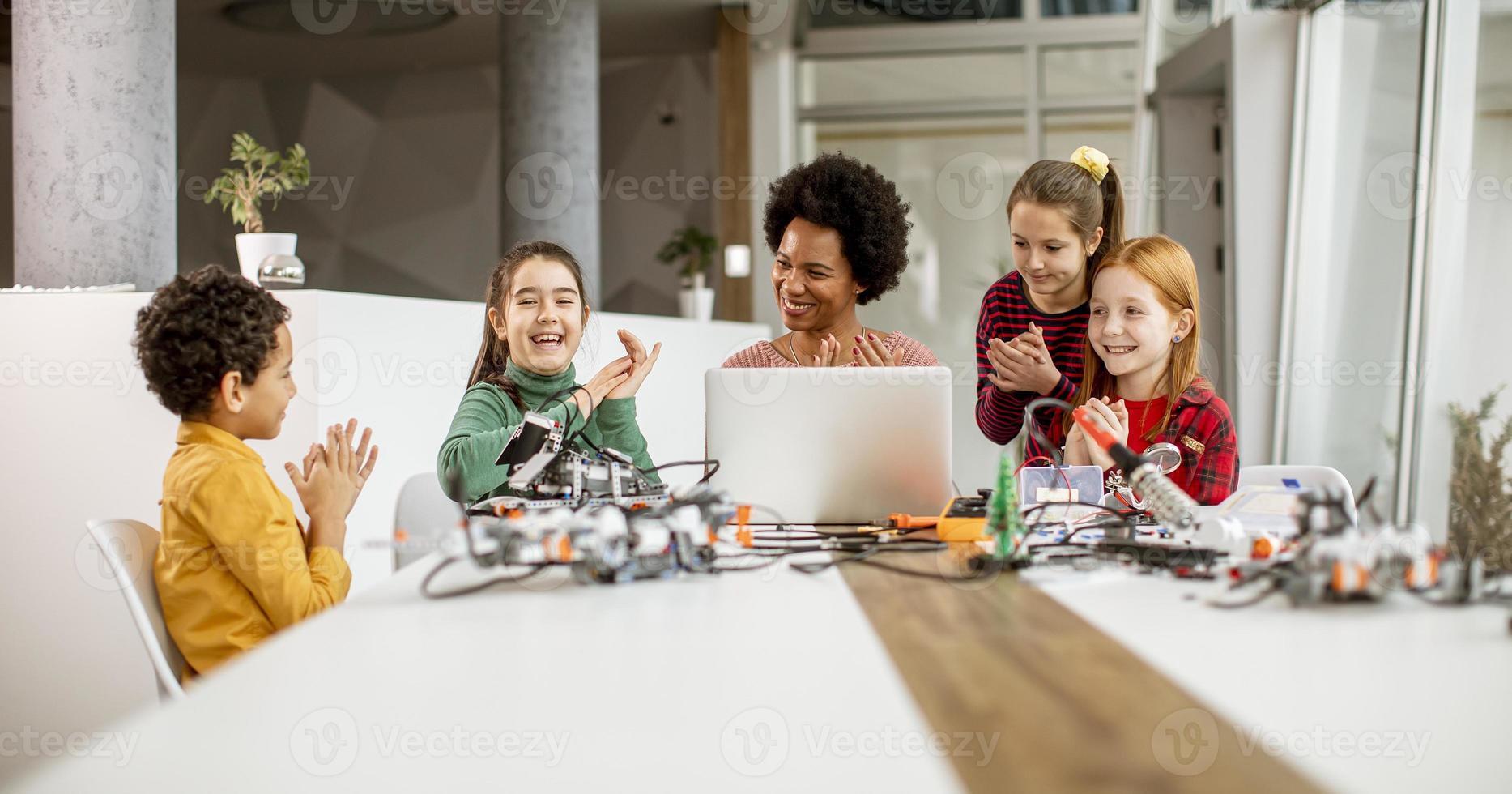 crianças felizes com sua professora de ciências afro-americana com laptop programando brinquedos elétricos e robôs na sala de aula de robótica foto