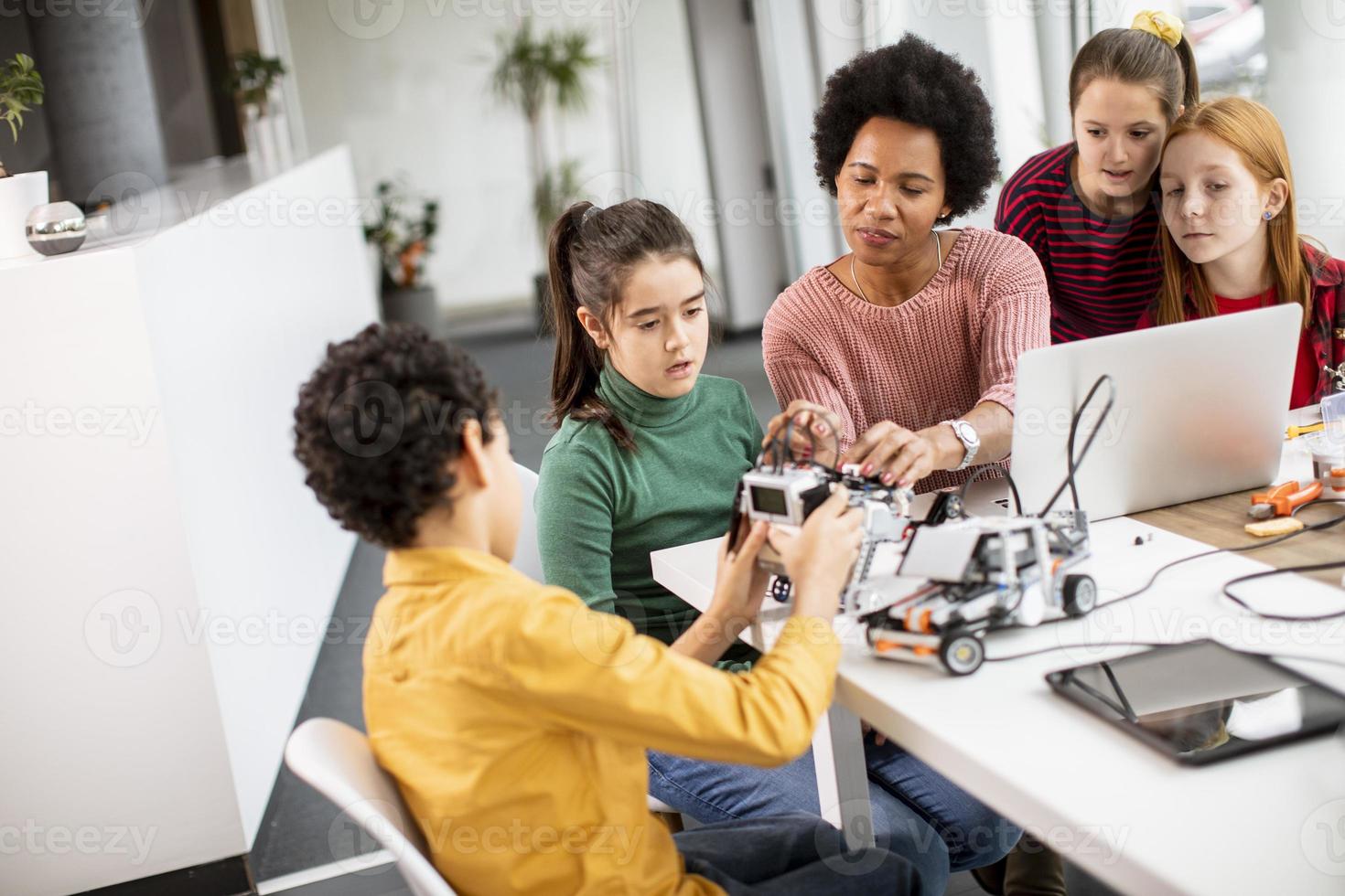 crianças felizes com sua professora de ciências afro-americana com laptop programando brinquedos elétricos e robôs na sala de aula de robótica foto