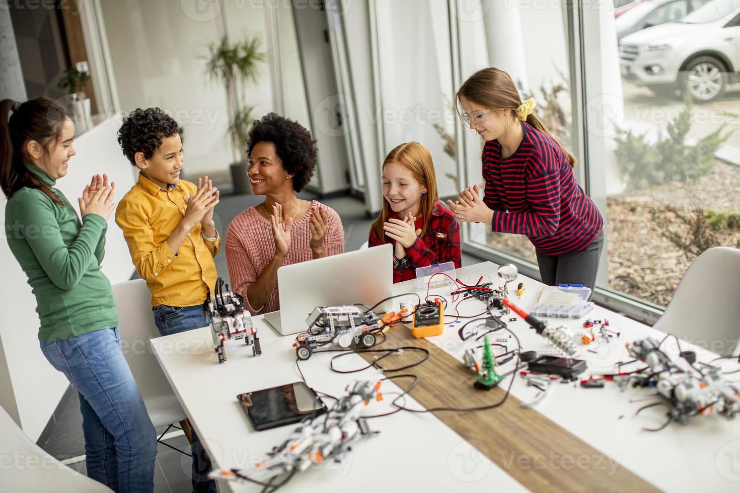 crianças felizes com sua professora de ciências afro-americana com laptop programando brinquedos elétricos e robôs na sala de aula de robótica foto