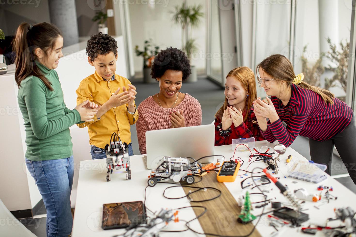 crianças felizes com sua professora de ciências afro-americana com laptop programando brinquedos elétricos e robôs na sala de aula de robótica foto