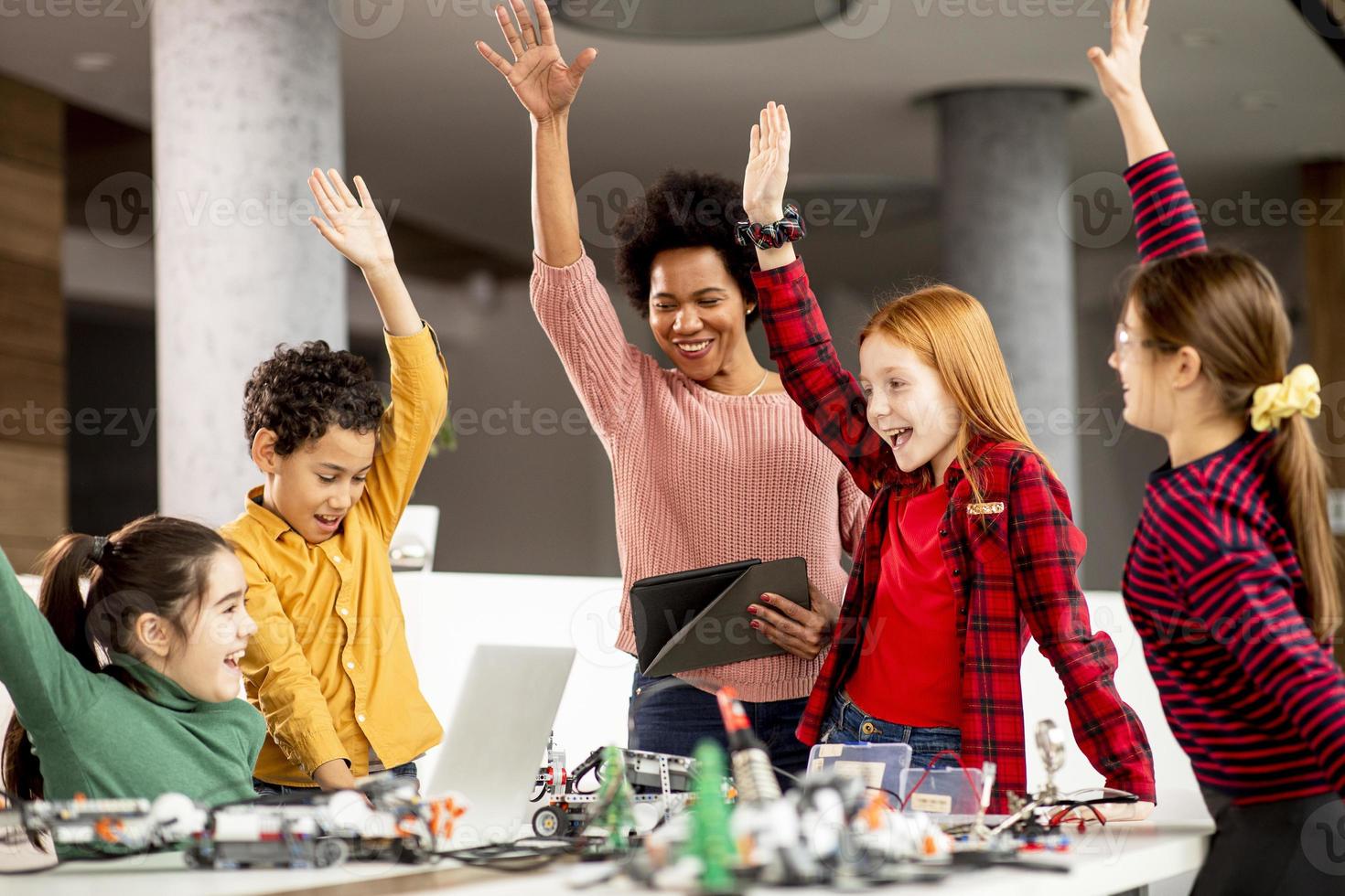 crianças felizes com sua professora de ciências afro-americana com laptop programando brinquedos elétricos e robôs na sala de aula de robótica foto