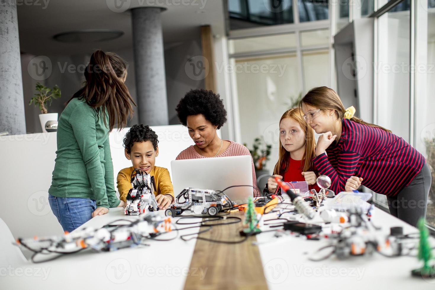 crianças felizes com sua professora de ciências afro-americana com laptop programando brinquedos elétricos e robôs na sala de aula de robótica foto