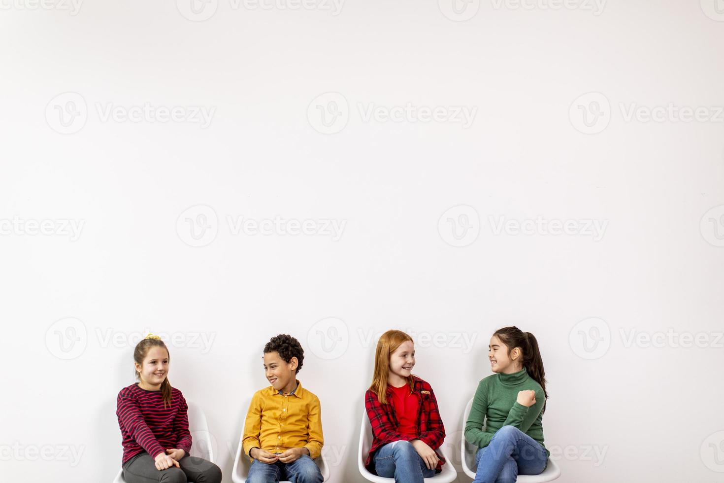 retrato de crianças fofas em jeans, sentadas em cadeiras contra uma parede branca foto