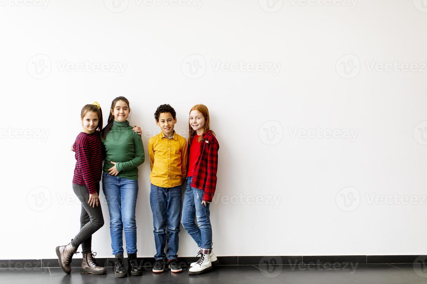 retrato de crianças fofas em jeans olhando para a câmera e sorrindo em pé contra uma parede branca foto