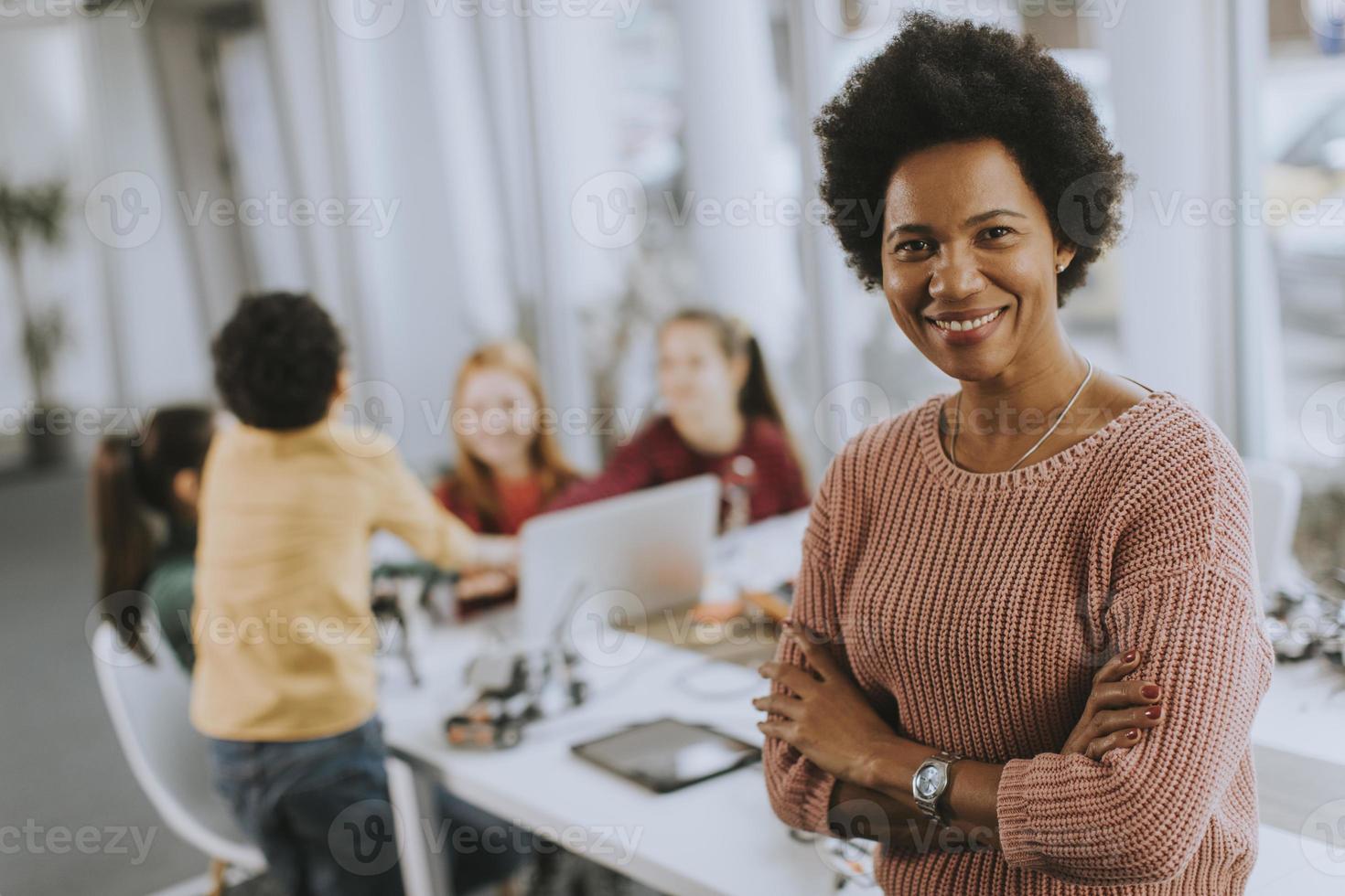 Professora afro-americana de ciências com um grupo de crianças programando brinquedos elétricos e robôs na sala de aula de robótica foto