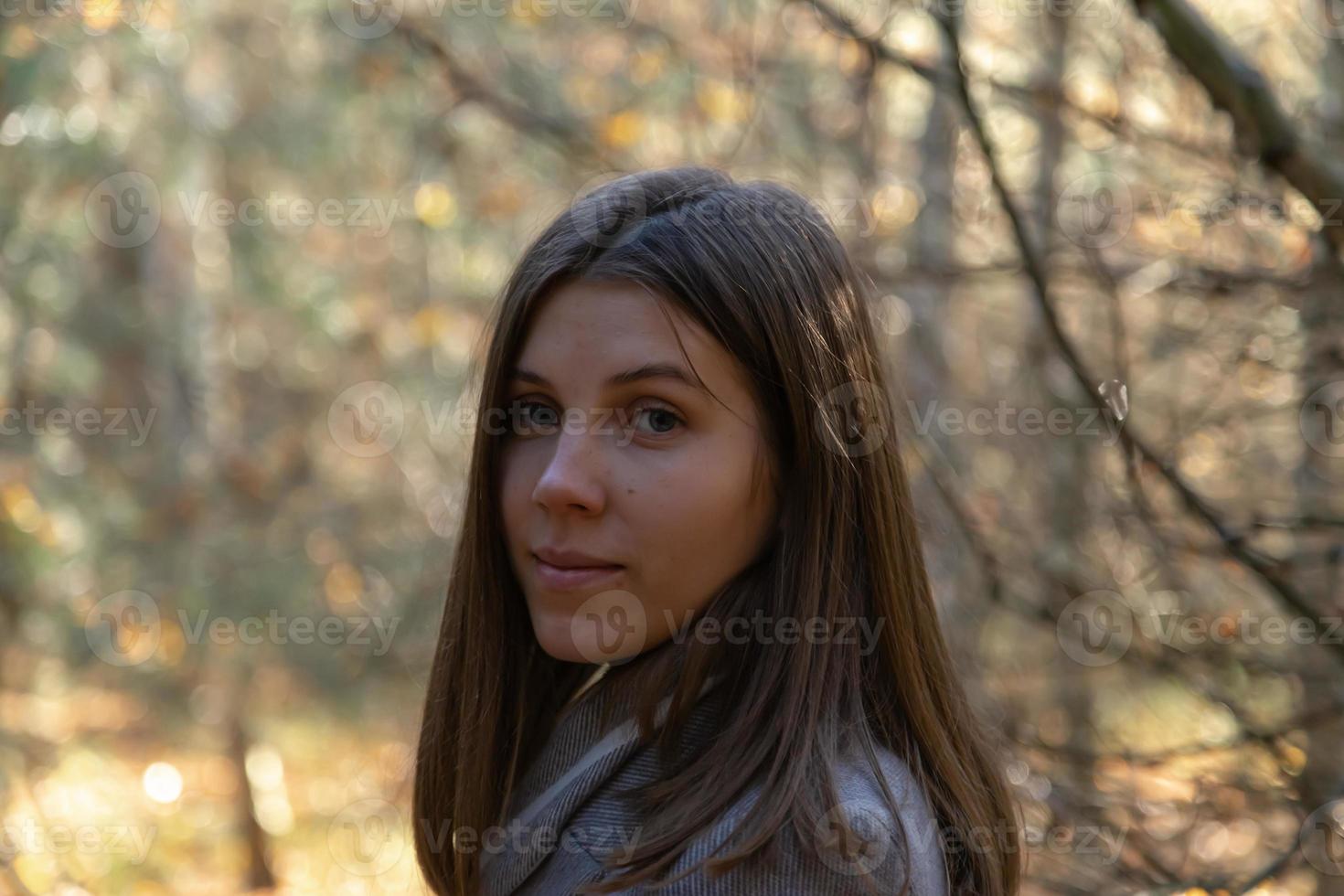 garota meio virada com um casaco cinza que fica na floresta em um dia ensolarado de outono foto