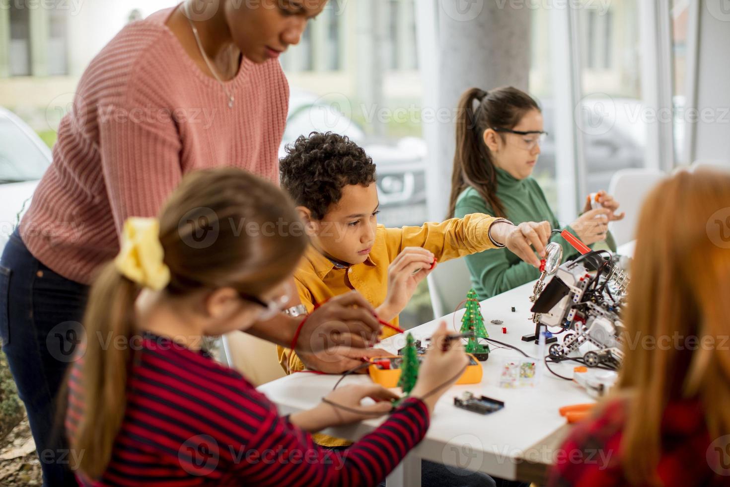 Professora afro-americana de ciências com um grupo de crianças programando brinquedos elétricos e robôs na sala de aula de robótica foto