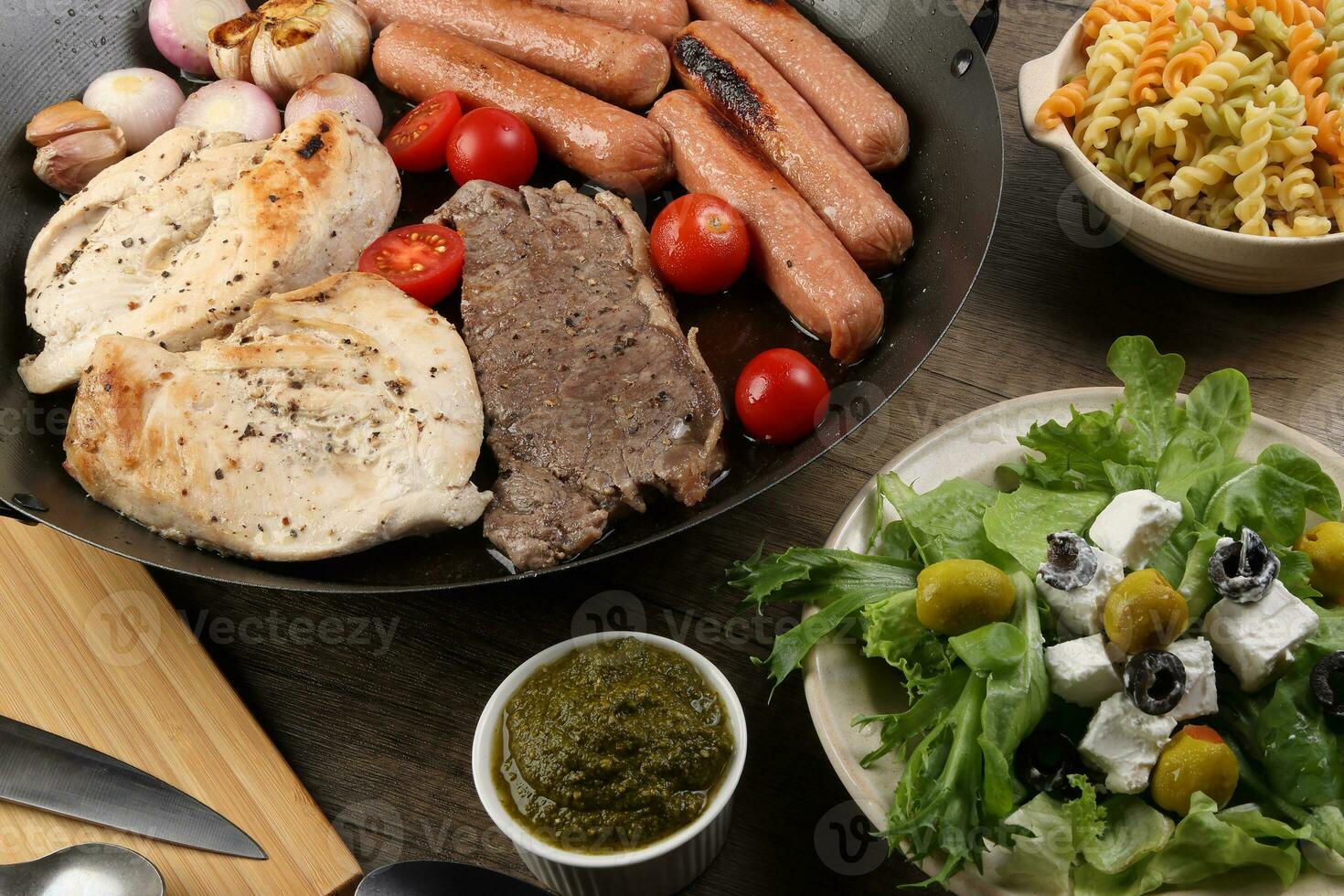 misturar Conheça frango carne bife linguiça tomate cebola alho fritar grade óleo metal fritar panela colorida massa verde salada com queijo Oliva hortelã manjericão pesto molho borda faca garfo colher rústico de madeira mesa foto