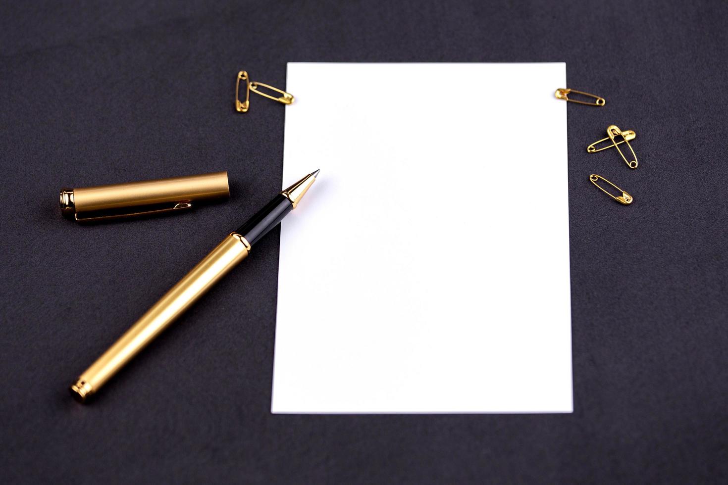 caneta, fita, clipes de papel e artigos de papelaria dourados em um fundo preto com uma folha de papel branca com espaço para cópia foto