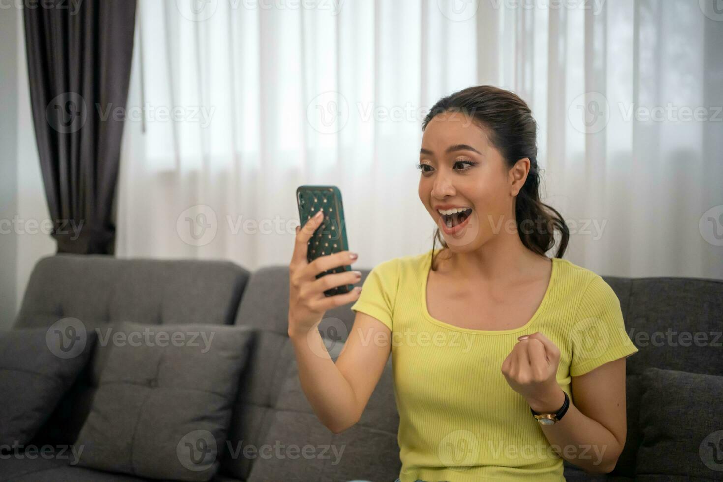 ásia mulher sentado em sofá às casa com Móvel telefone e sentindo-me animado e feliz. foto