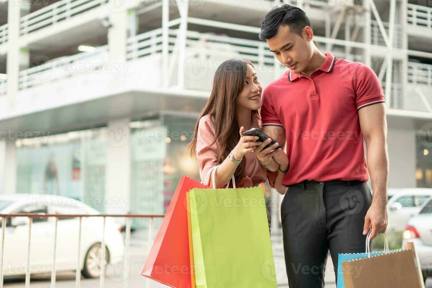 casal jovem feliz de compradores caminhando na rua comercial em direção e segurando sacolas coloridas na mão e usam um smartphone para promoção de cheques. conceito de venda e compras de sexta-feira negra foto