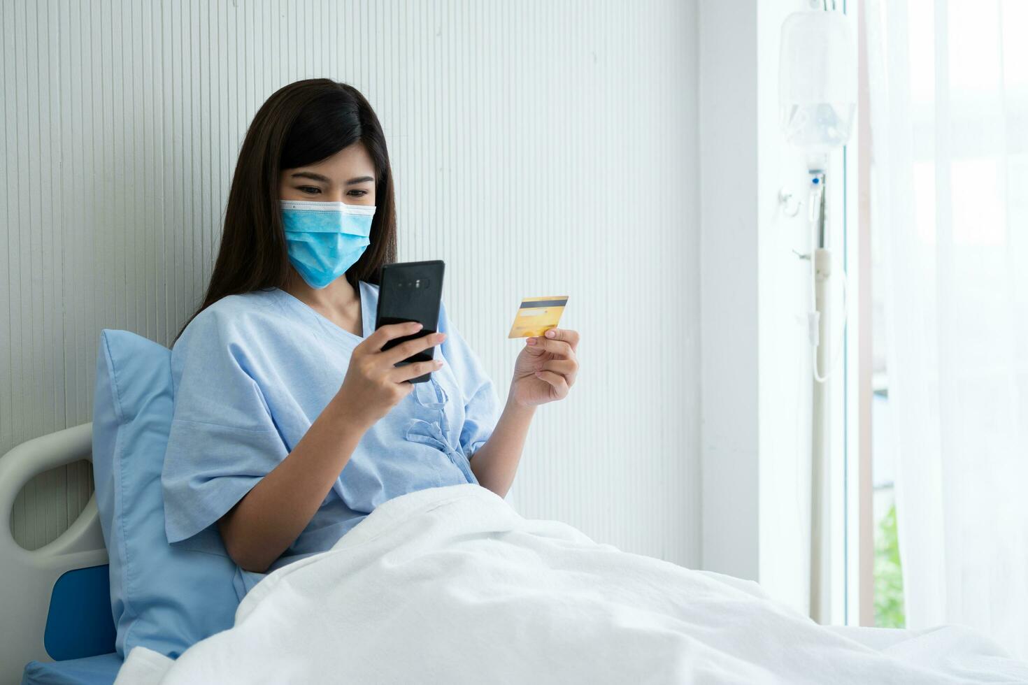 feliz ásia mulher vestindo uma médico mascarar e segurando zombar acima crédito seguro cartão e Smartphone dentro uma hospital cama para Verifica saúde seguro elegibilidade. seguro política de banco, Forma de pagamento médico foto