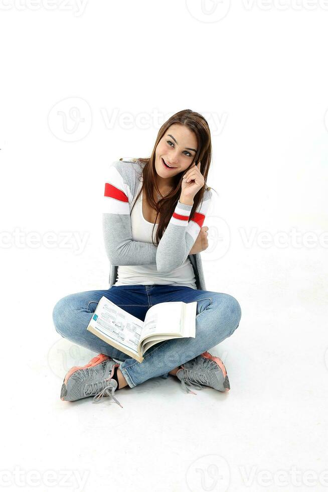 Yong ásia juventude menina dentro casual vestir sentar em chão livro em colo mão em bochecha expressão engraçado espirituoso em branco fundo foto