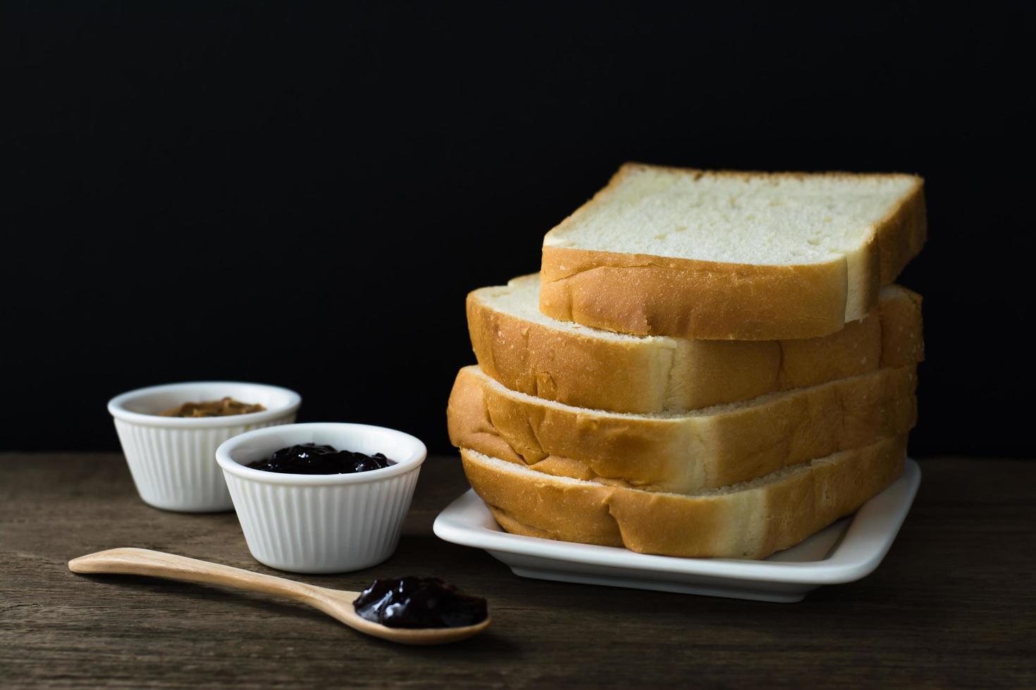 fatias de pão torrado em um único prato branco na mesa de madeira com fundo preto e geleia de groselha preta no copo pequeno e na colher de pau foto