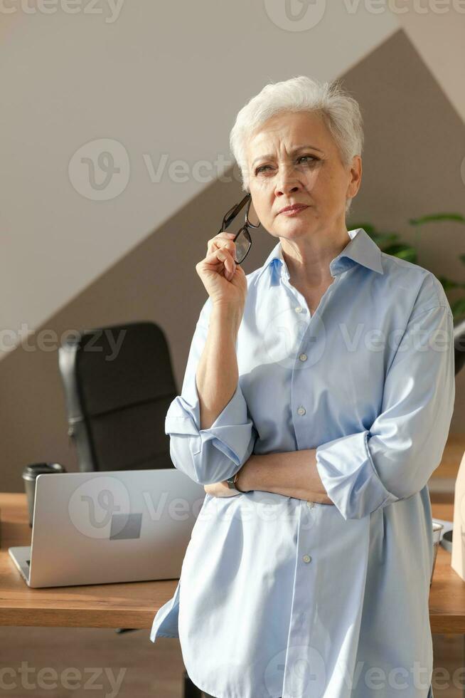 retrato do confiante à moda europeu meio envelhecido Senior mulher às ambiente de trabalho. à moda Mais velho maduro anos 60 cinzento cabelos senhora empresária dentro moderno escritório. patrão líder professor profissional trabalhador. foto