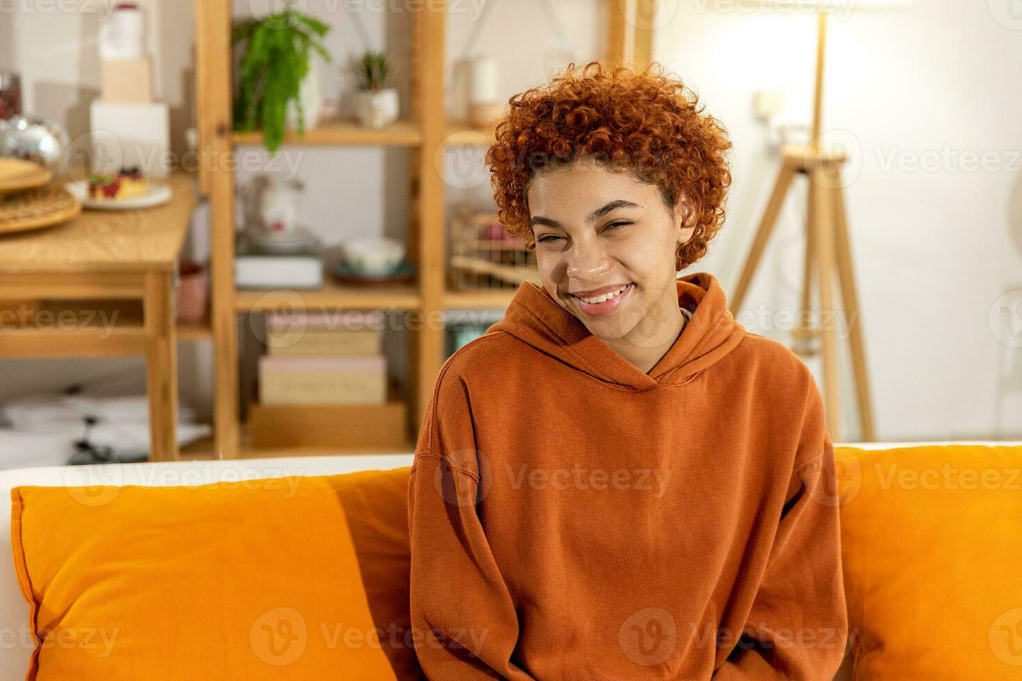 linda garota afro-americana com penteado afro sorrindo sentado no sofá em casa interior. jovem africana com cabelos cacheados rindo. liberdade felicidade despreocupado conceito de pessoas felizes. foto