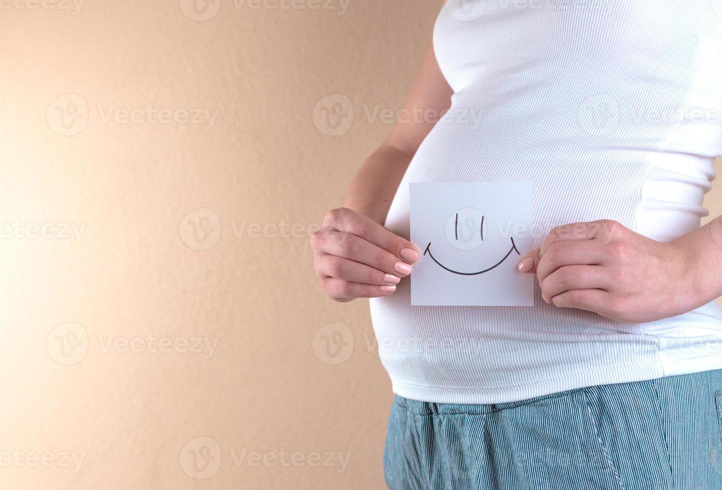 uma visão aproximada da barriga de uma mulher grávida segurando um pedaço de papel com uma carinha sorridente foto