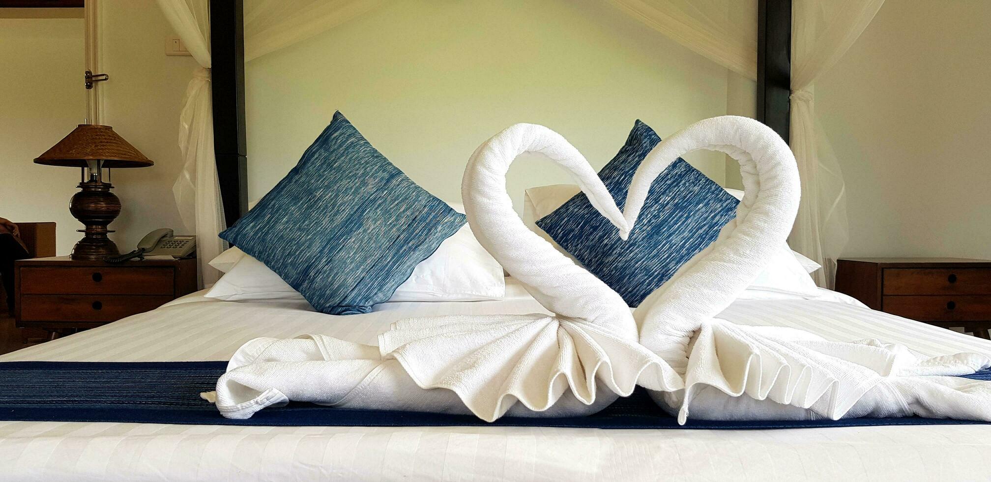 lindo branco toalha guardada dentro dois cisne forma ou coração com azul travesseiro e parede fundo em quarto dentro hotel. lindo Projeto e idéia para decoração quarto e morador conceito foto