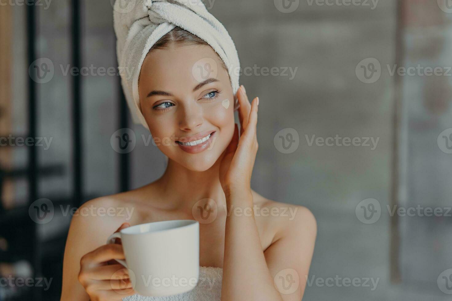 bonita mulher aplica-se face creme, mínimo inventar, embrulhado dentro banho toalha, goza café às lar. beleza, relaxamento conceito. foto