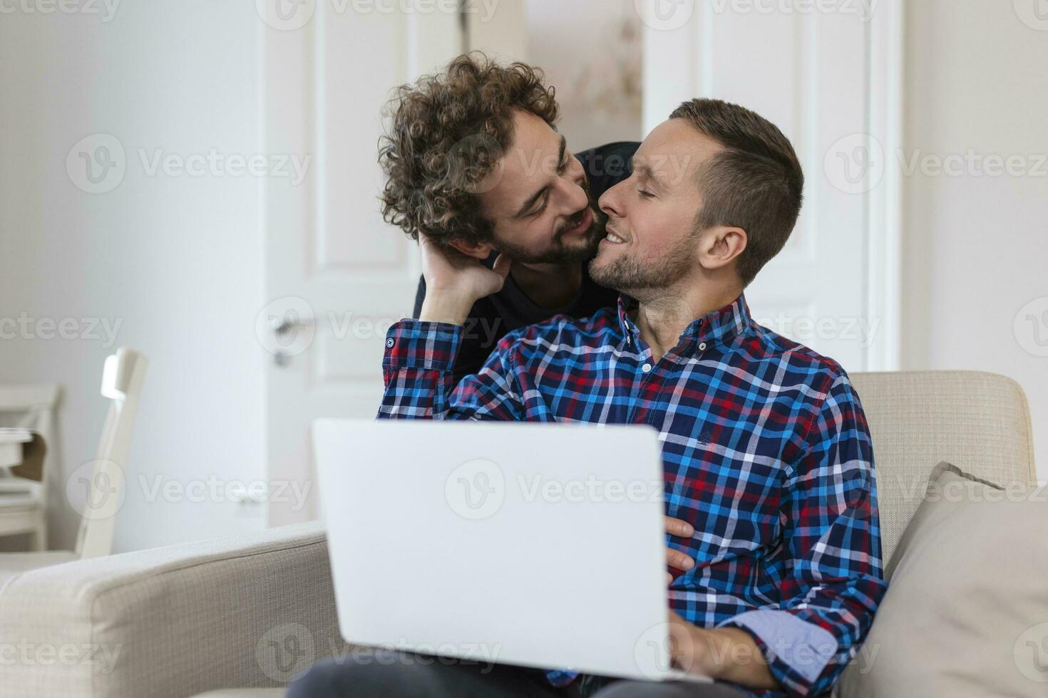 adorável gay casal rindo juntos enquanto sentado dentro seus vivo quarto às lar. dois romântico jovem masculino amantes tendo Diversão surfar a Internet dentro de casa. jovem gay casal vivo junto. foto