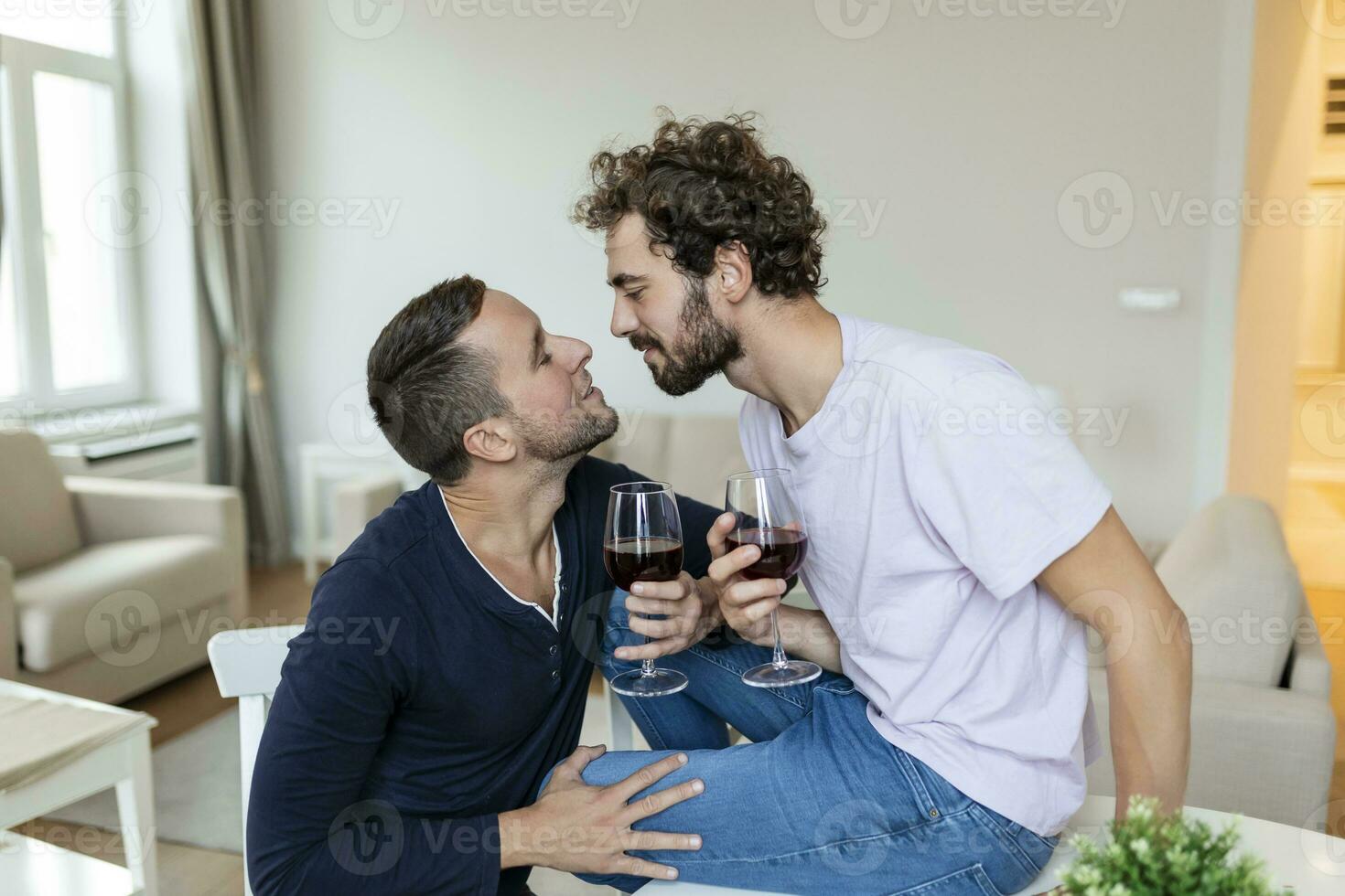 lgbtq casal abraçando cada de outros e bebendo vinho dentro de casa. dois romântico jovem masculino amantes olhando às cada de outros enquanto sentado juntos dentro seus vivo sala. jovem gay casal ser romântico às lar. foto