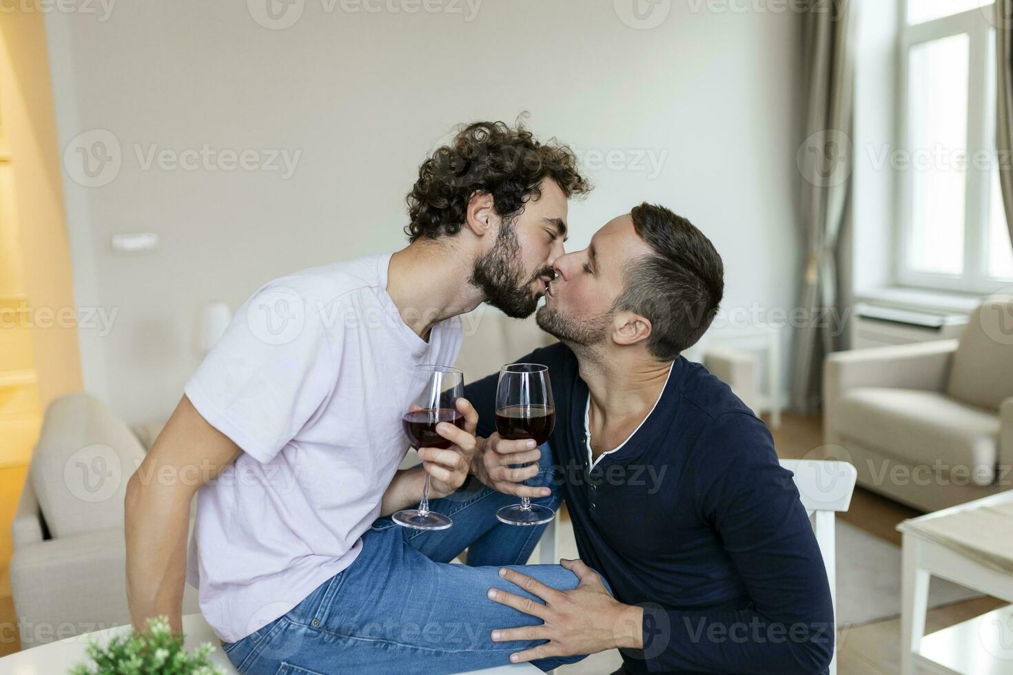 lgbtq casal abraçando cada de outros e bebendo vinho dentro de casa. dois romântico jovem masculino amantes olhando às cada de outros enquanto sentado juntos dentro seus vivo sala. jovem gay casal ser romântico às lar. foto