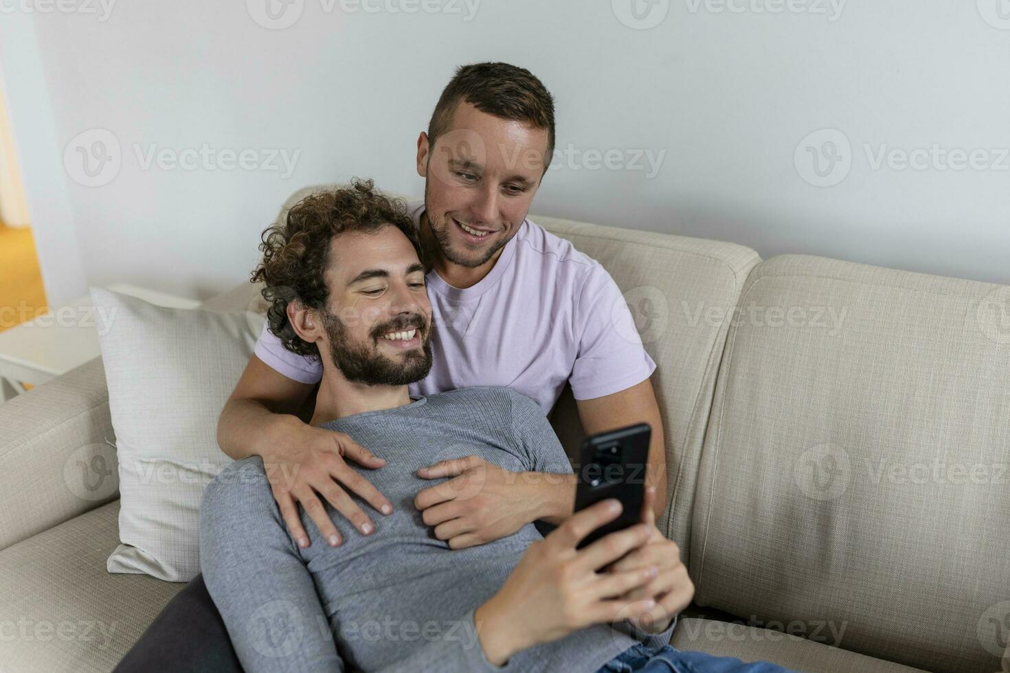 fofa jovem gay casal vídeo chamando seus amigos dentro seus vivo quarto às lar. dois masculino amantes sorridente alegremente enquanto cumprimento seus amigos em uma Smartphone. jovem gay casal sentado junto. foto