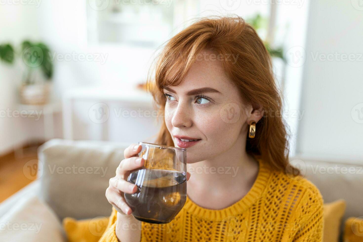 feliz jovem mulher sorridente enquanto segurando uma vidro do água às lar. mulher em vivo quarto sofá relaxado e sorridente enquanto bebendo vidro do água. saúde benefícios do bebendo o suficiente água foto