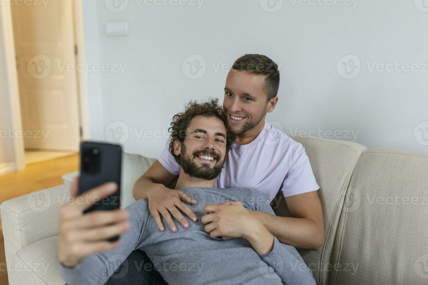 fofa jovem gay casal vídeo chamando seus amigos dentro seus vivo quarto às lar. dois masculino amantes sorridente alegremente enquanto cumprimento seus amigos em uma Smartphone. jovem gay casal sentado junto. foto