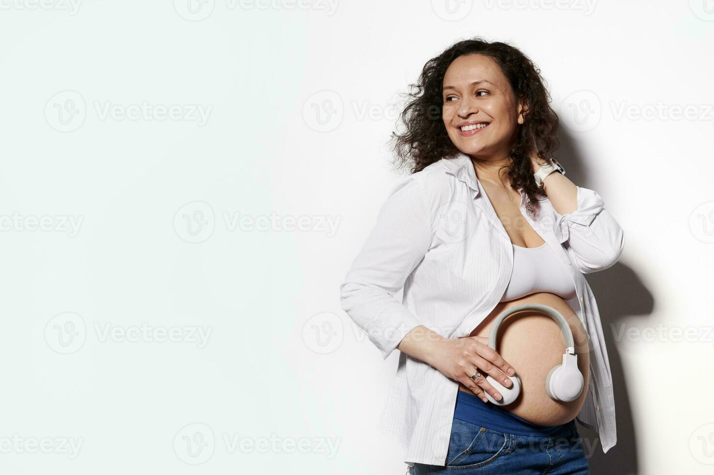 sorridente relaxado grávida mulher, futuro mãe com barriga coloca fones de ouvido em barriga para bebê para ouço para música. gravidez foto