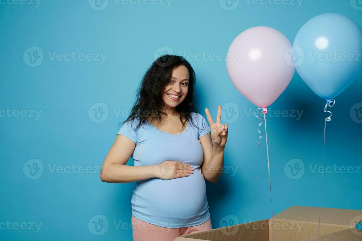 grávida mulher sorrisos golpes barriga mostra dois dedos às Câmera, sentindo-me positivo emoções esperando gêmeos. gênero festa foto