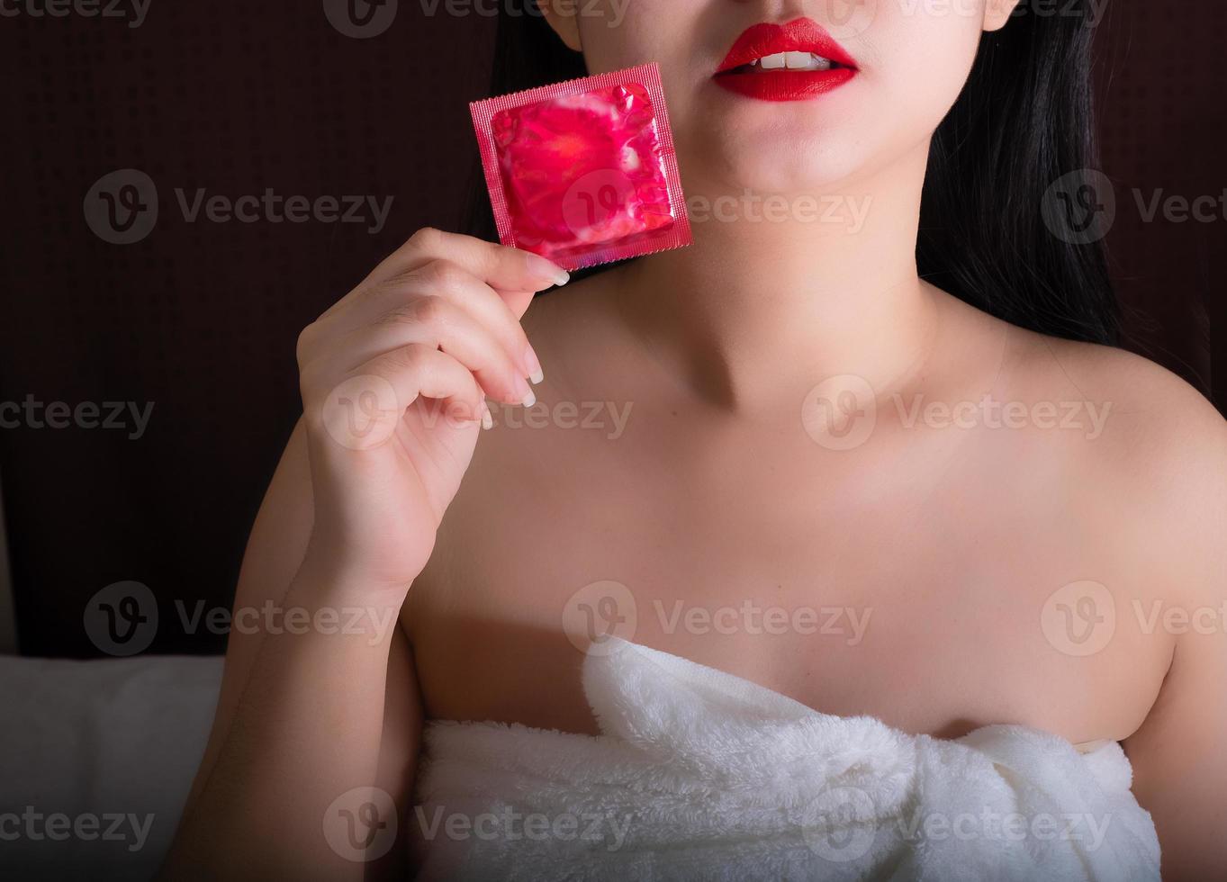 preservativo vermelho na mão em primeiro plano para a mulher bonita com uma toalha branca deitada na cama foto