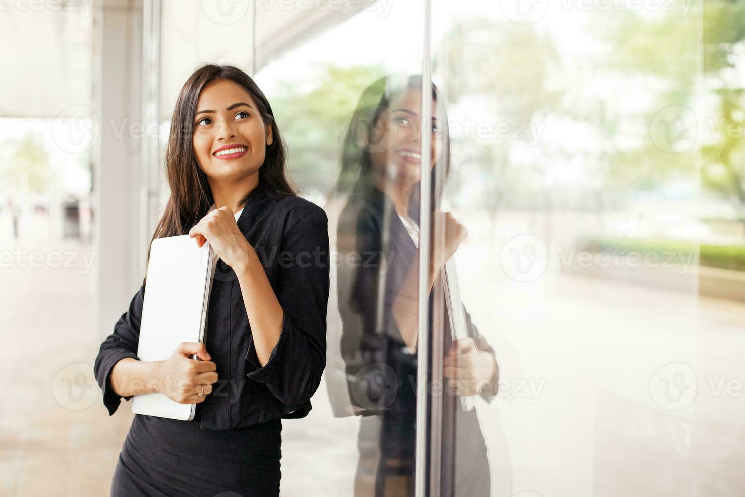 uma lindo profissional indiano mulher vestindo uma terno em pé segurando uma computador portátil e sorridente de uma vidro janela foto