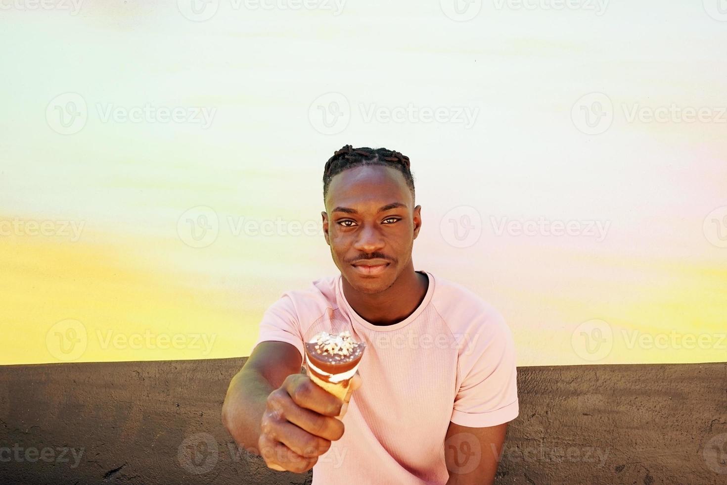 jovem negro bonito usa uma camisa rosa segura e come uma casquinha de sorvete no verão em uma parede pintada como um nascer do sol ou dia ensolarado foto