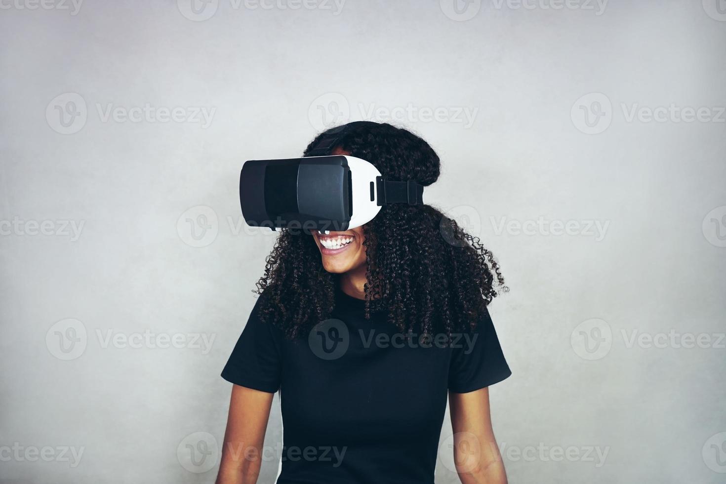 uma bela jovem negra com cabelo afro encaracolado usa fone de ouvido de realidade virtual vr e joga videogame enquanto sorri no estúdio com fundo cinza foto