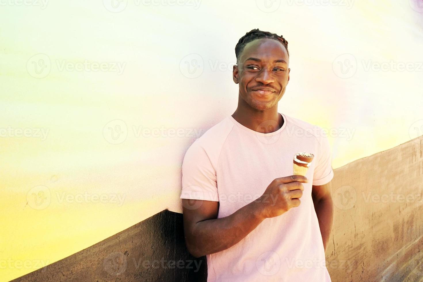 jovem negro bonito usa uma camisa rosa segura e come uma casquinha de sorvete no verão em uma parede pintada como um nascer do sol ou dia ensolarado foto