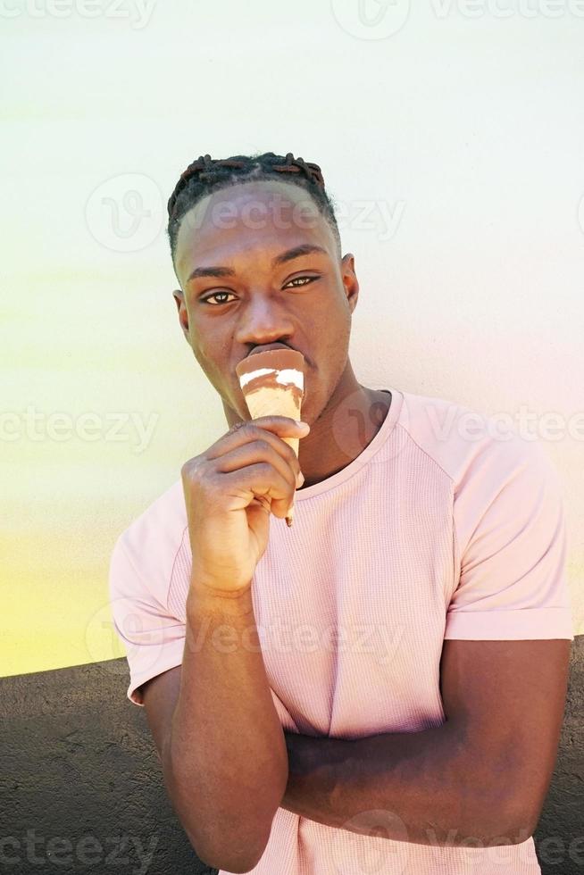 jovem negro bonito usa uma camisa rosa segura e come uma casquinha de sorvete no verão em uma parede pintada como um nascer do sol ou dia ensolarado foto