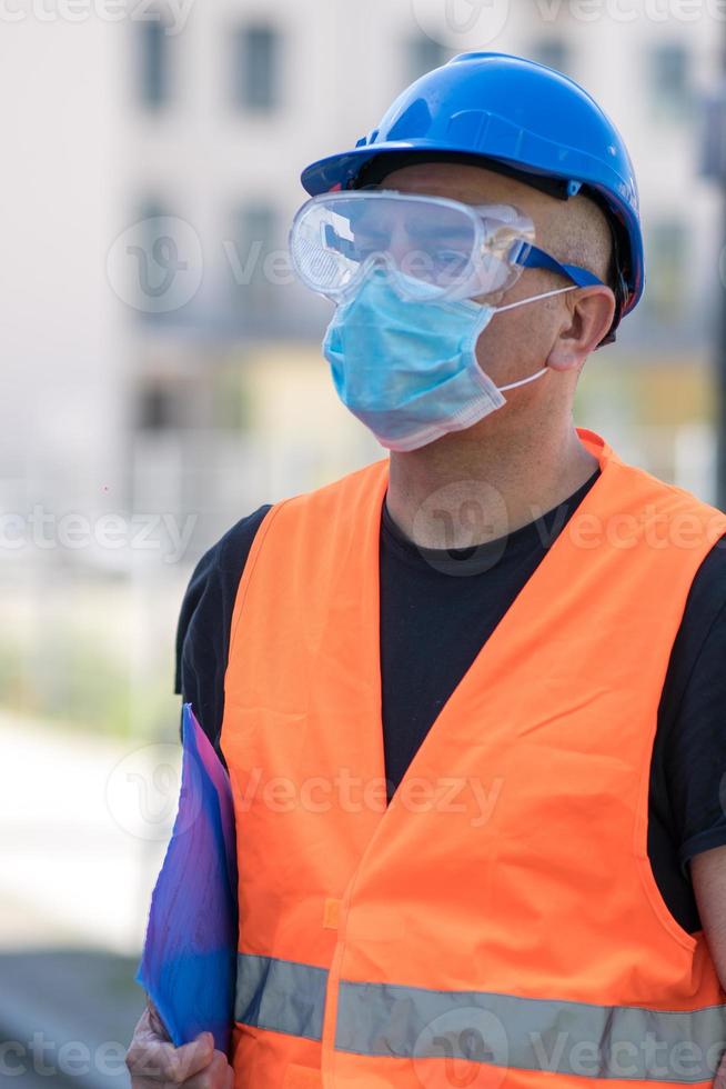 trabalhador da construção civil usando capacete azul, colete reflexivo e máscara cirúrgica protetora foto