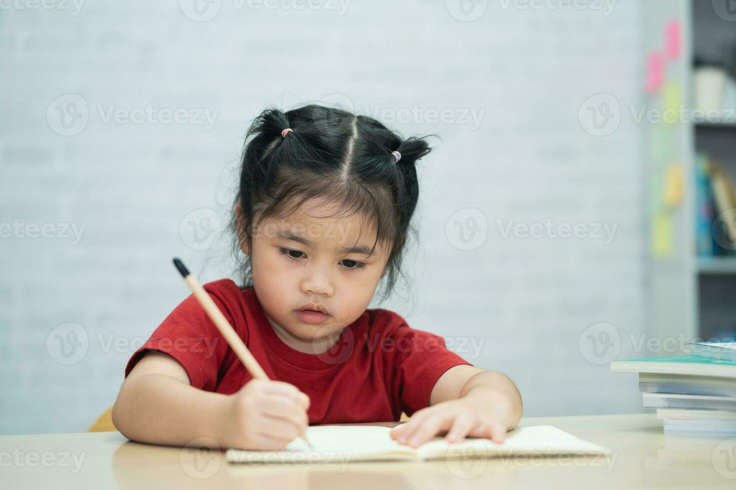 ásia bebê menina vestindo uma vermelho camisa escrever notas dentro caderno e lendo livro para estude conectados em madeira mesa escrivaninha dentro vivo quarto às lar. Educação Aprendendo conectados a partir de casa conceito. foto