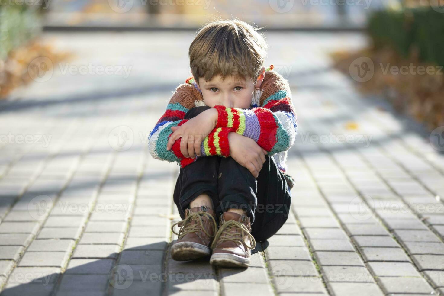 triste pequeno Garoto senta em a beco dentro a parque. a criança colocar dele cabeça em dele joelho. foto