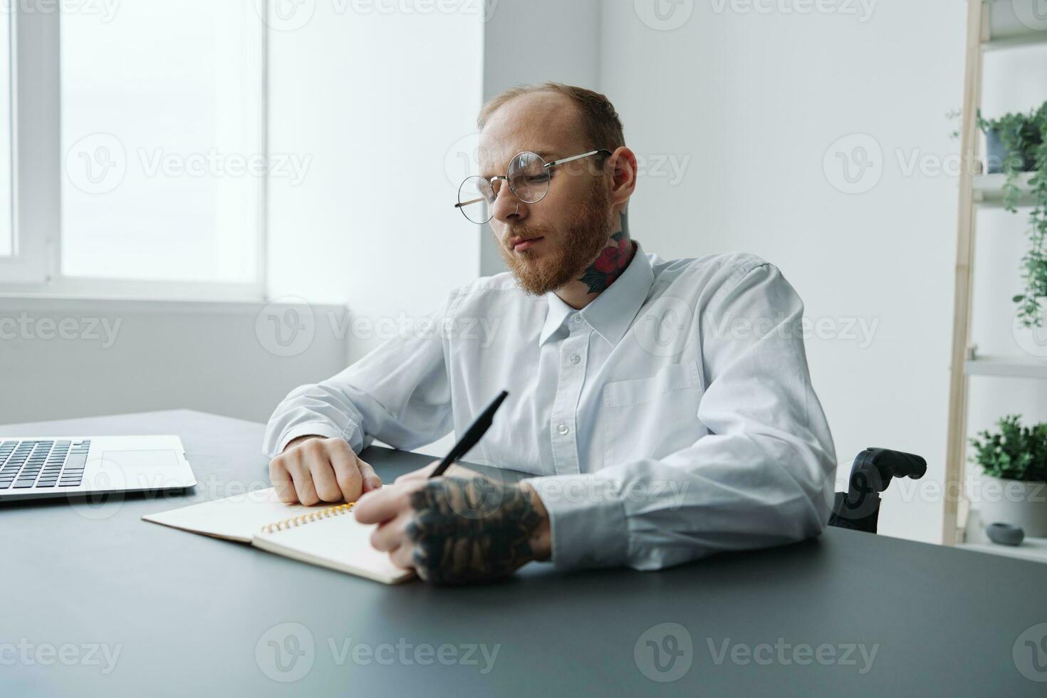 uma homem dentro uma cadeira de rodas, uma homem de negocios dentro a escritório trabalho às uma computador portátil, escrevendo baixa uma plano dentro uma caderno, Consideração, integração para dentro sociedade, a conceito do trabalhando uma pessoa com deficiências foto