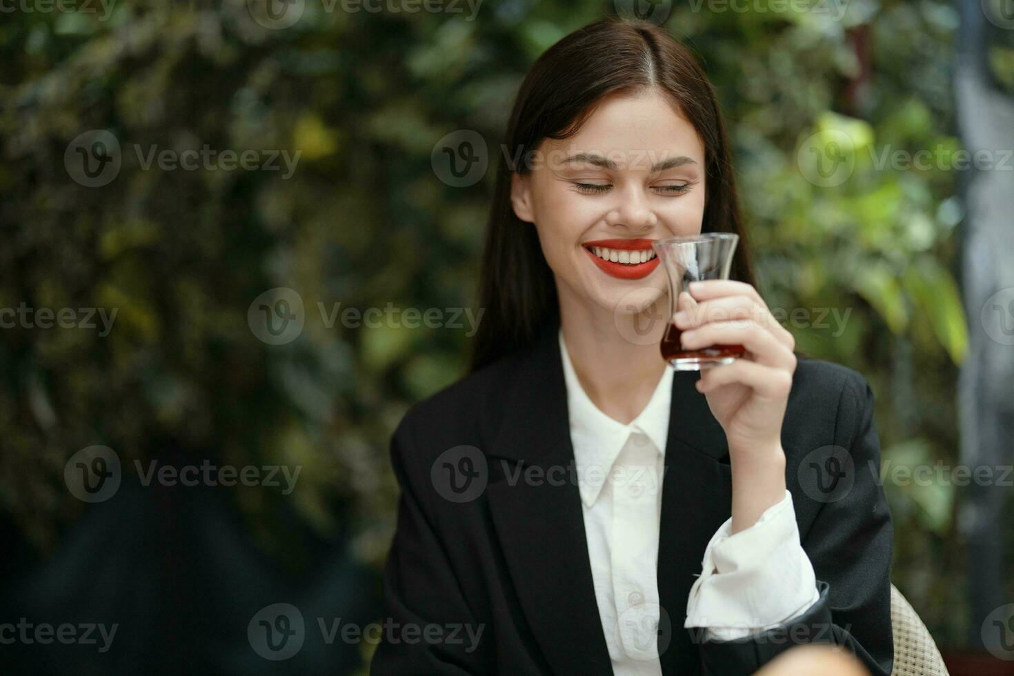 mulher com vermelho lábios sorrir com dentes bebidas chá dentro uma cafeteria a partir de uma turco vidro caneca dentro uma branco camisa e Preto moda Jaqueta em a rua, verão viagem, período de férias dentro a cidade foto