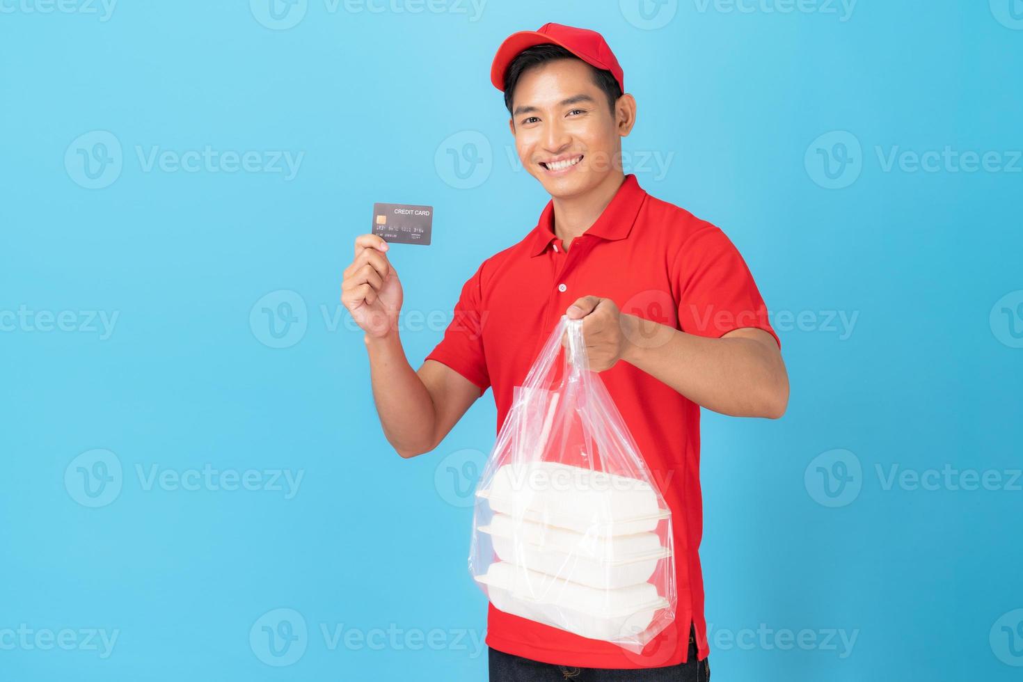 Empregado entregador sorridente com boné vermelho camisa em branco uniforme permanente com cartão de crédito dando pedido de comida foto