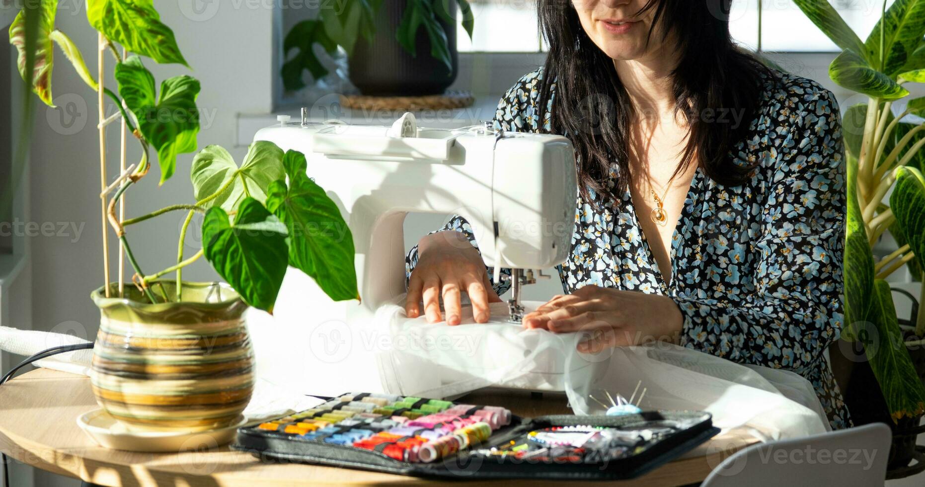 uma mulher costura tule em a elétrico de costura máquina dentro uma branco moderno interior do uma casa com ampla janelas, casa plantas. conforto dentro a casa, uma dona de casa passatempo foto