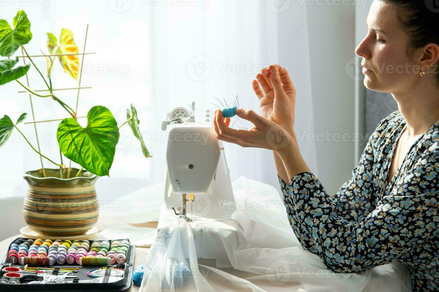 uma mulher costura tule em a elétrico de costura máquina dentro uma branco moderno interior do uma casa com ampla janelas, casa plantas. conforto dentro a casa, uma dona de casa passatempo foto