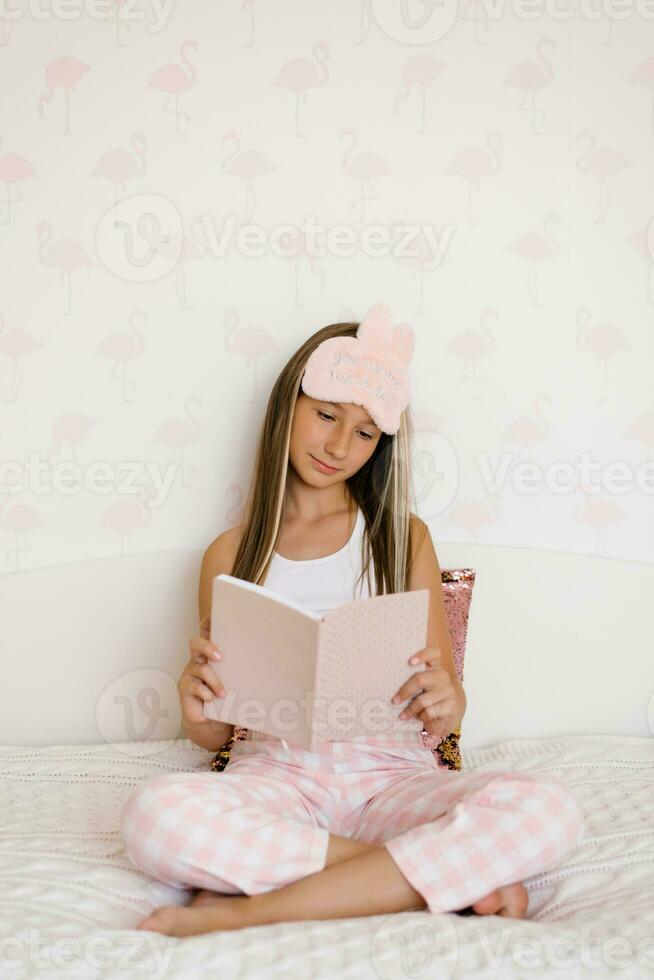 Adolescência menina é sentado em a cama dentro dela pijamas dentro a manhã e lendo uma livro. acolhedor final de semana às casa foto