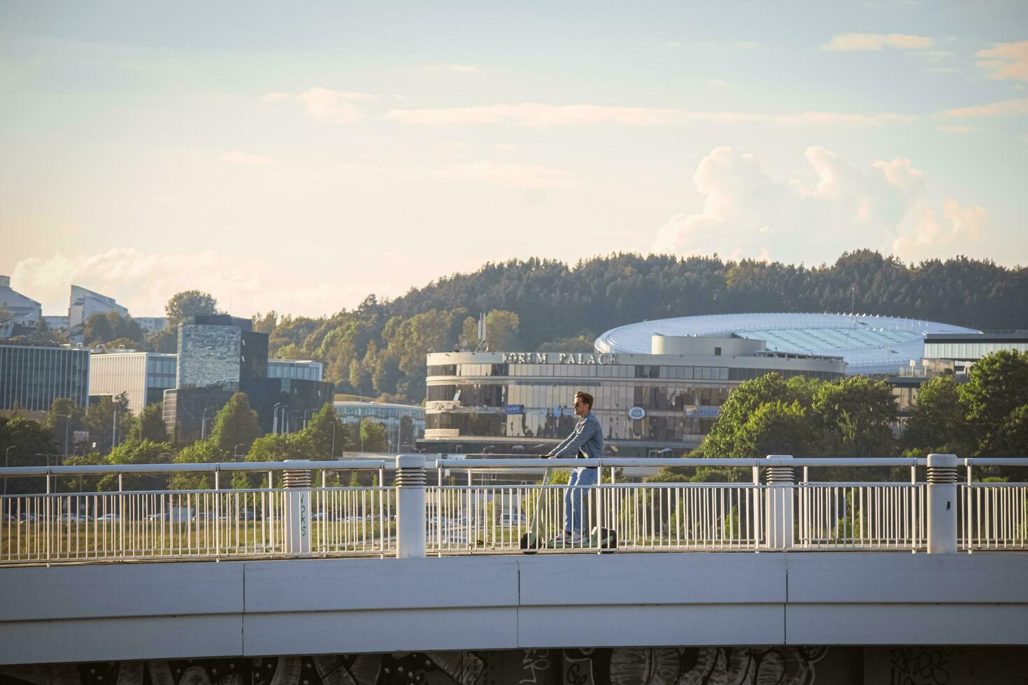 Vilnius, Lituânia 03 06 2022 jovem homem em lambreta dirigindo ao longo uma branco ponte sobre rio com verde árvores e escritório edifícios em fundo foto