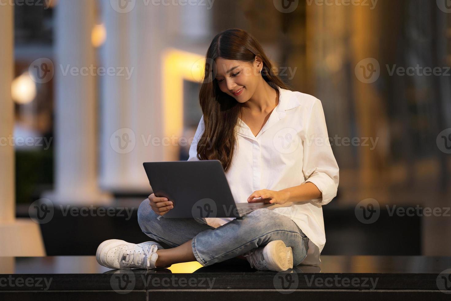 jovem asiática com uma carinha sorridente usando laptop em uma cidade à noite foto