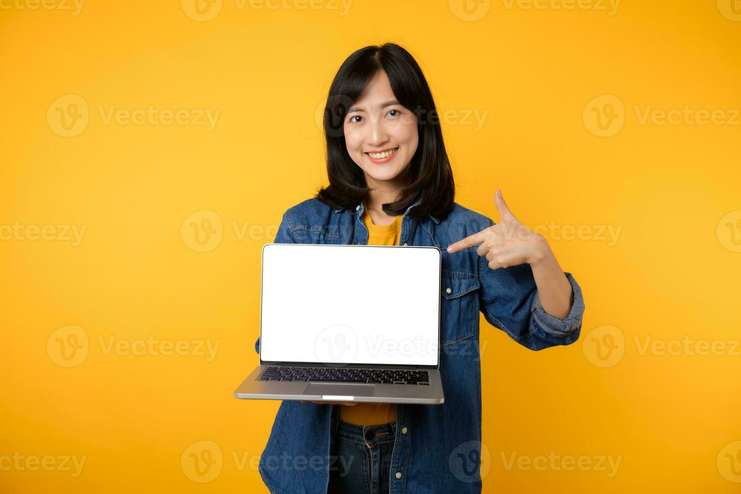 retrato jovem feliz mulher vestindo amarelo camiseta e jeans camisa segurando computador portátil e ponto dedo para tela isolado em amarelo estúdio fundo. o negócio tecnologia inscrição comunicação conceito. foto